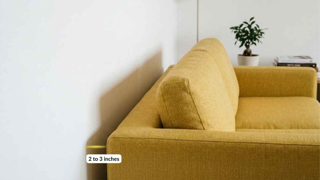 Close-up side angle of a yellow fabric sofa pulled exactly 2 inches away from a white wall. A thin visible shadow line runs along the wall where the gap creates perceived depth. Warm soft light illuminates the shadow gap. Demonstrates the shadow gap principle for making a small room look bigger without renovation.